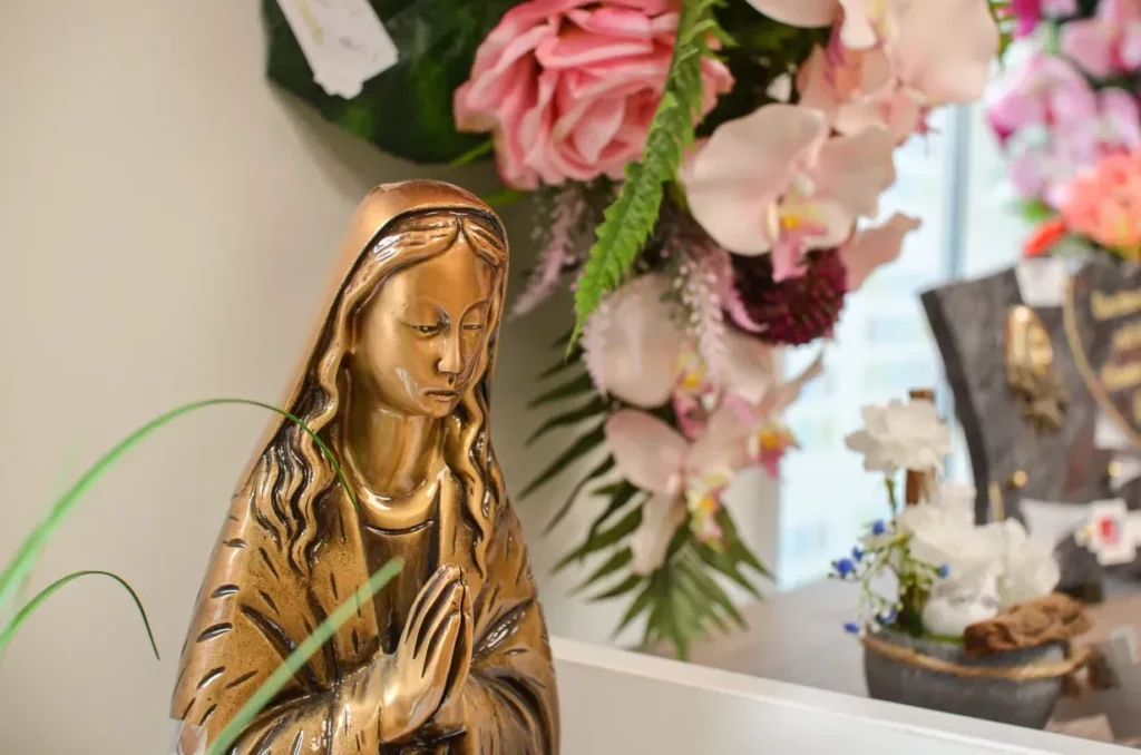 Statuette religieuse de la Vierge Marie en prière devant une gerbe de fleurs artificielles.